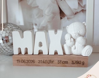 May include: A white decorative set featuring the name MAX in block letters next to a textured teddy bear figure. These rest on a rectangular wooden base engraved with the text 19.06.2026, 21:41 Uhr, 51 cm, and 3.780 g, alongside a silver disco ball.