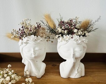 May include: Two white matte ceramic head planters shaped like faces with closed eyes. One figure rests a hand on its cheek, while the other holds a finger to its lips in a shushing gesture. Both are adorned with floral crowns filled with dried flowers.