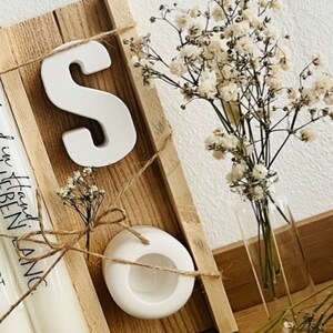 May include: A wooden shelf with a white ceramic letter "S", a white ceramic candle holder, and a bouquet of white baby's breath in a clear glass vase.