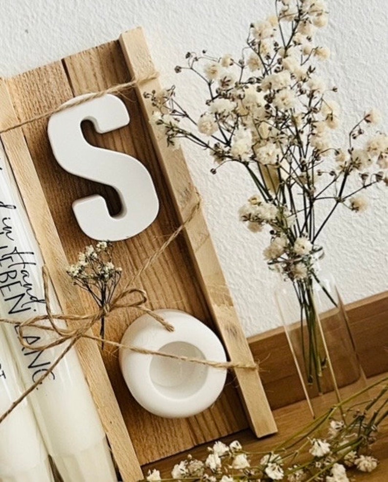 May include: A wooden shelf with a white ceramic letter "S", a white ceramic candle holder, and a bouquet of white baby's breath in a clear glass vase.