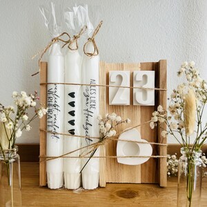 May include: Three white candles with the text "Alles Liebe Hochzeits" and "nicht mehr verliebt, verheiratet" on them. The candles are tied together with twine and are sitting on a wooden board with the numbers "23" on it. There is a small white heart-shaped candle holder on the board.
