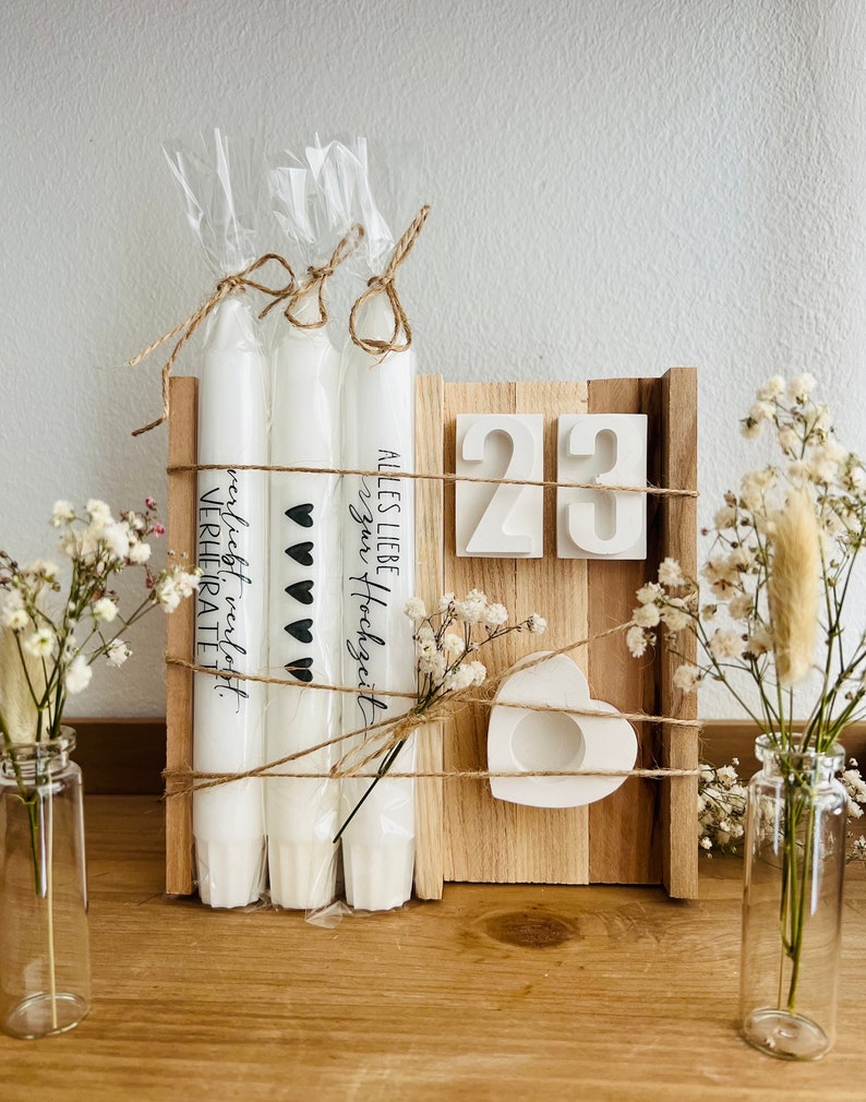 May include: Three white candles with the text "Alles Liebe Hochzeits" and "nicht mehr verliebt, verheiratet" on them. The candles are tied together with twine and are sitting on a wooden board with the numbers "23" on it. There is a small white heart-shaped candle holder on the board.