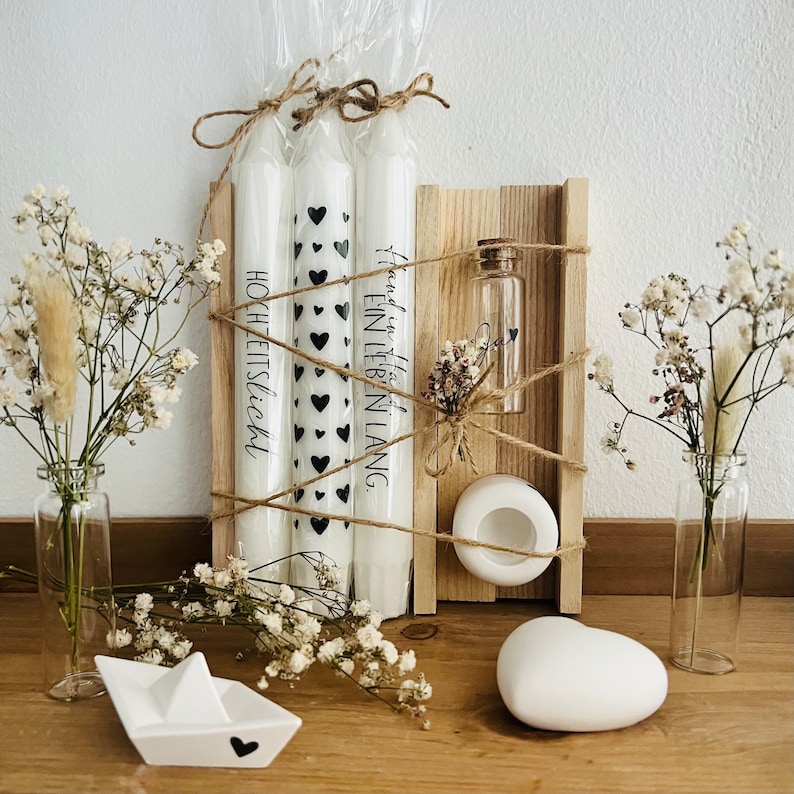 May include: A wooden box with three white candles, a small glass bottle with a cork top, a white heart-shaped object, and a white ceramic dish with a hole in the center. The candles are wrapped in clear plastic and have black hearts printed on them. The candles have the text "Hochzeitslicht" and "Happy in this life long" printed on them.