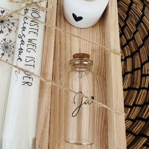 May include: A wooden box with a white candle holder, a small glass bottle with a cork top, and a white scroll with the text "Der h&ouml;chste Weg ist der  einesames" in black lettering.