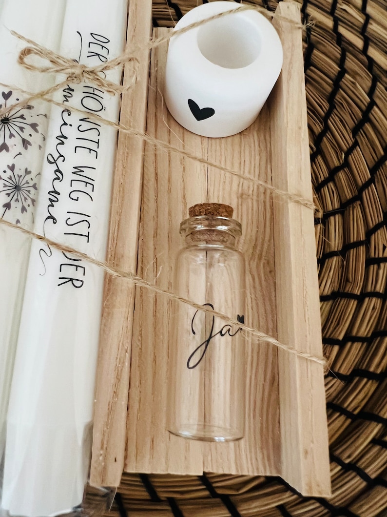 May include: A wooden box with a white candle holder, a small glass bottle with a cork top, and a white scroll with the text "Der h&ouml;chste Weg ist der  einesames" in black lettering.