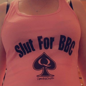 May include: Pink tank top with the text "Slut For BBC" in black lettering. A black spade with a queen inside is below the text. The text "Spades & Castle" is below the spade.