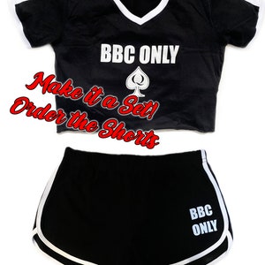 Pu&ograve; includere: Top corto nero e pantaloncini abbinati con bordini bianchi. Il top e i pantaloncini hanno il testo "BBC ONLY" e un simbolo di picche stampato su di essi. Il testo "Make it a Set! Order the Shorts" &egrave; scritto in rosso.