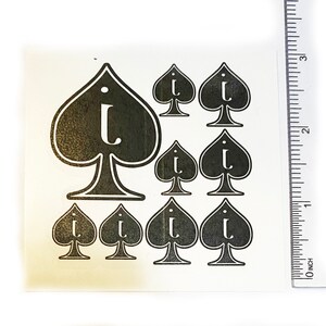 May include: A sheet of black and white spade-shaped stickers, each featuring the letter "i". The stickers are of varying sizes, with a large spade at the top and smaller ones below. A ruler, marked in inches, is visible on the right.