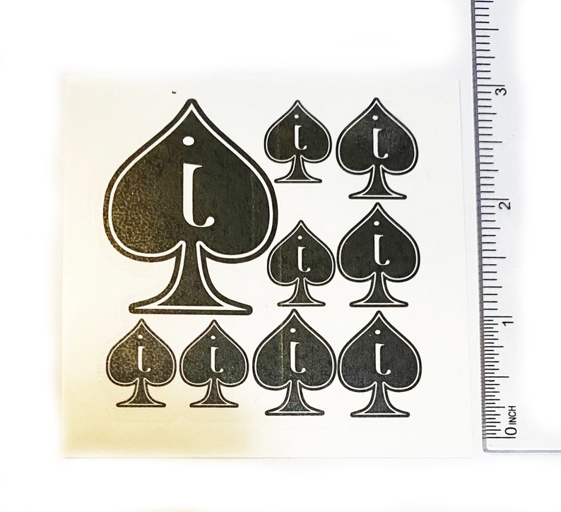 May include: A sheet of black and white spade-shaped stickers, each featuring the letter "i". The stickers are of varying sizes, with a large spade at the top and smaller ones below. A ruler, marked in inches, is visible on the right.