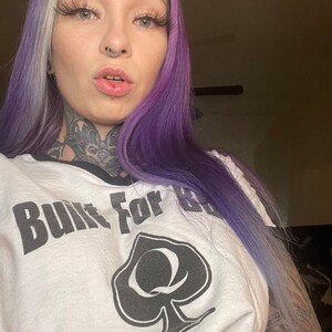K&ouml;nnte beinhalten: Wei&szlig;es T-Shirt mit einem schwarzen Pik und dem Text "Built For" auf der Vorderseite. Die Person, die das Shirt tr&auml;gt, hat lila und blonde Haare.