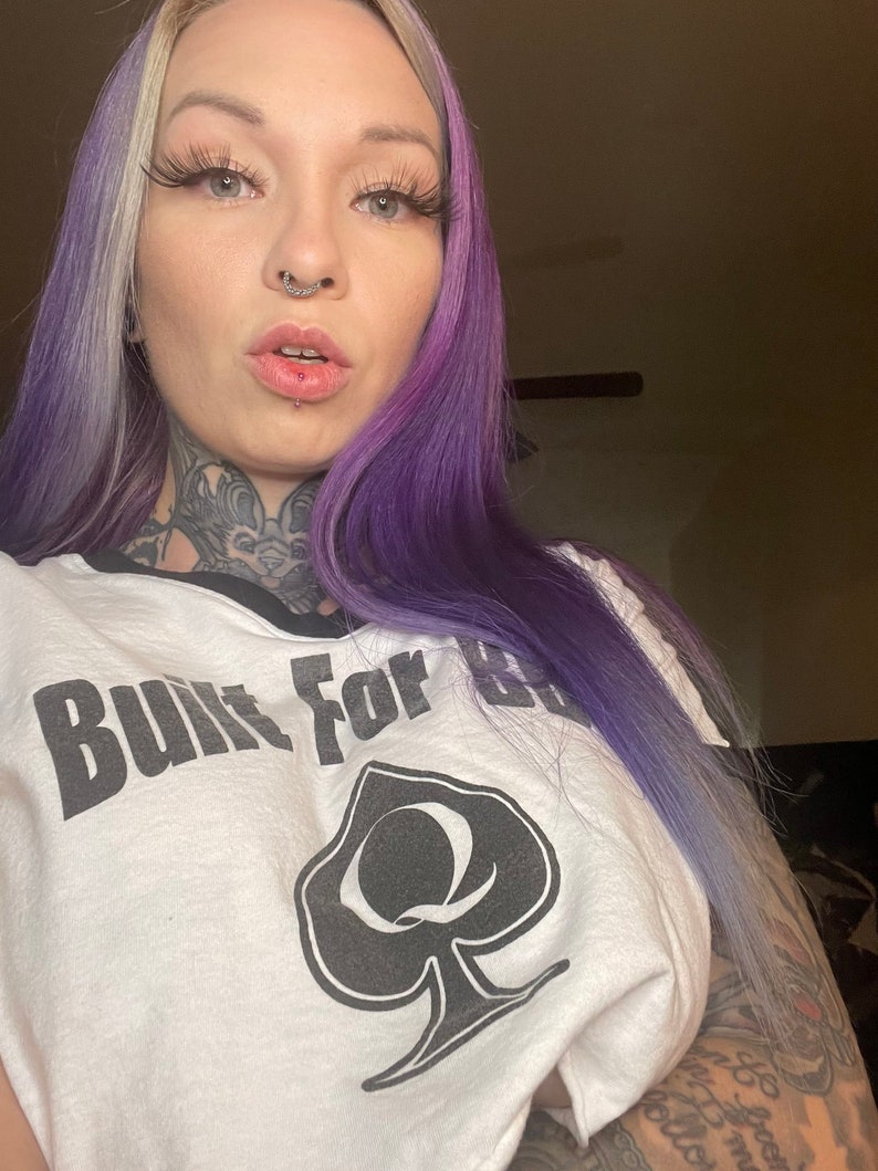 K&ouml;nnte beinhalten: Wei&szlig;es T-Shirt mit einem schwarzen Pik und dem Text "Built For" auf der Vorderseite. Die Person, die das Shirt tr&auml;gt, hat lila und blonde Haare.