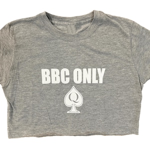 Pu&ograve; includere: Una maglietta grigia a taglio corto con il testo "BBC ONLY" in lettere bianche e un simbolo di picche bianco con una "Q" al suo interno.