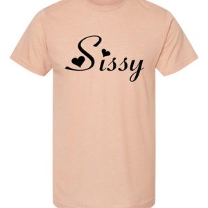 May include: A peach-colored t-shirt with the word "Sissy" in black script, featuring heart accents. The short-sleeved shirt is made of a soft material.