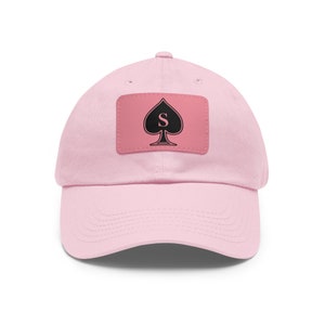 Puede incluir: Una gorra de béisbol rosa con un parche negro que presenta una pica con la letra 'S' en su interior.