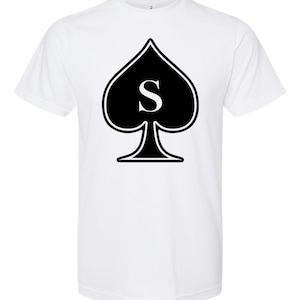 May include: White t-shirt featuring a black spade symbol with a white "S" in the center. The spade design is outlined in black. The shirt has short sleeves and a crew neck.