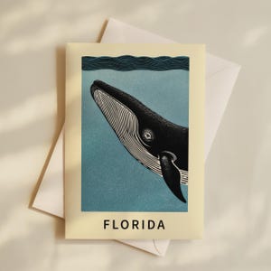 May include: A cream-coloured greeting card with a stylised illustration of a whale swimming underwater. The whale is black with white markings, set against a blue background. The word "FLORIDA" is printed in black at the bottom.