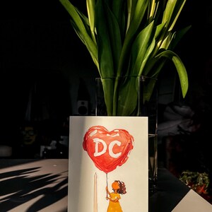 Watercolor illustration of a girl holding a red heart-shaped balloon with "DC" written on it, standing near The Washington Monument. The charming postcard captures the spirit of Washington, DC, with a dreamy, whimsical touch. Perfect for DC lovers, travel enthusiasts, and postcard collectors, this 4x6 design makes a unique souvenir or gift. Printed on premium cardstock with a matte finish, it’s ideal for mailing, framing, or adding to your collection.