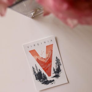 Virginia postcard featuring the letter "V" artistically intertwined with lush forests and majestic mountain peaks. A perfect 4x6" Virginia souvenir celebrating the state’s natural beauty. Printed on premium matte cardstock, ideal for nature lovers, hikers, and adventure seekers.