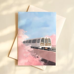 May include: A greeting card featuring a train traveling on a bridge, surrounded by pink clouds, under a blue sky. The train is white with brown accents. The card is inside a white envelope.