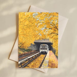 May include: Greeting card featuring a train emerging from a tunnel, set against a backdrop of vibrant yellow autumn foliage. The train is white with a gray roof, and the scene is rendered in a painterly style. The card is paired with a cream-colored envelope.