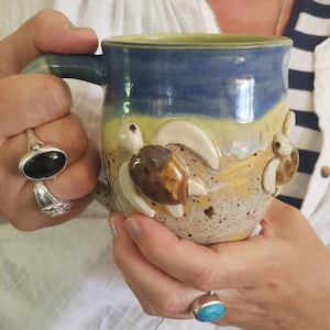 May include: A blue and yellow ceramic mug with three turtle figurines attached to the side. The mug is speckled with brown and white spots.