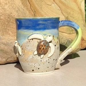 May include: A ceramic mug with a blue, yellow and white glaze. The mug features three sculpted sea turtles, one of which is brown with a white shell. The mug has a handle with a yellow accent.