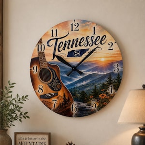 Op de afbeelding: Een ronde wandklok met een berglandschap bij zonsondergang, een akoestische gitaar en het woord Tennessee in script. De wijzerplaat toont zwarte cijfers van 1 tot 12, zwarte wijzers en een klein icoon van de vlag van de staat Tennessee.