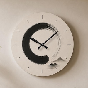 May include: A round wall clock with a cream-colored face featuring a black ink-wash style circular brushstroke and a small mountain landscape illustration. The clock has black hour and minute hands and simple black line markers for the hours.