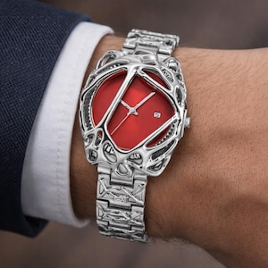 Könnte beinhalten: Eine silberfarbene Armbanduhr mit einem leuchtend roten Zifferblatt. Die Uhr hat ein kompliziertes, durchbrochenes Design um das Zifferblatt und am Armband. Die Uhr wird am Handgelenk getragen, mit einem Anzugsakko und einem weißen Hemd im Hintergrund.