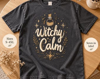 Könnte beinhalten: Ein anthrazitfarbenes Kurzarm-T-Shirt mit einem Grafikdruck aus Zaubertrankflasche, Sternen und Blättern sowie dem Schriftzug "Witchy Calm" in cremefarbener Schreibschrift. Das T-Shirt hat einen Rundhalsausschnitt und eine Vintage-Waschung.