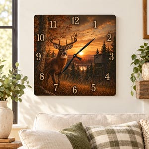 Puede incluir: Un reloj de pared cuadrado con una escena impresa de un ciervo de pie en un bosque al atardecer. La imagen incluye un ciervo, pinos, un puesto de caza y un cielo dorado. La esfera del reloj tiene manecillas negras y números grandes.