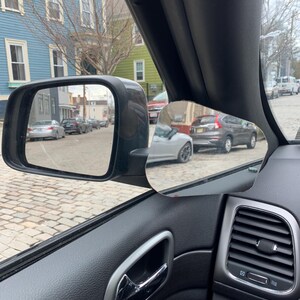 May include: A black car's interior with a side view mirror showing a street scene with parked cars and buildings. The car's door handle and air vent are visible.