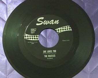 May include: A green vinyl record with a white label. The label has the text "Swan" in a cursive font. The record is titled "She Loves You" by "The Beatles". The record label also includes the text "Don't Drop Out" and "Gil Music Corp. BMI".
