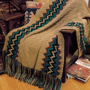 Könnte beinhalten: Eine gehäkelte Wolldecke mit einem geometrischen Muster in Braun, Türkis und Schwarz. Die Decke hat einen Fransenrand und ist über einen Holzstuhl gelegt.