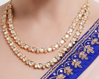 May include: A woman wearing a gold necklace with white stones and gold earrings with white stones. She is wearing a light brown top and a blue and gold patterned sari.