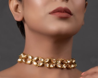 May include: A gold-toned choker necklace with a floral design. The necklace features a double-strand design with alternating leaf-shaped elements. The leaves are filled with a white material and accented with green and gold details. The jewelry is worn on a person.
