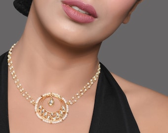 May include: A woman wearing a gold-toned necklace with a circular pendant. The pendant is adorned with small white pearls and clear gemstones. The woman has pink eyeshadow and lipstick. The background is a solid gray.