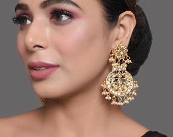 May include: A pair of gold earrings with white pearl accents. The earrings feature a circular design with intricate details and a dangling pearl drop.