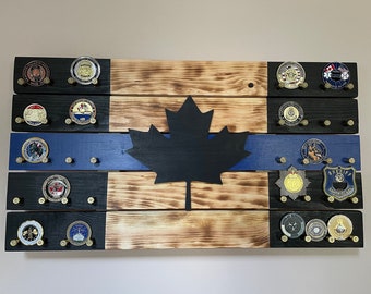 Op de afbeelding: Een houten wanddisplay met een zwart silhouet van een Canadees esdoornblad. Het display heeft een blauwe streep en is versierd met verschillende gouden en zilveren badges.
