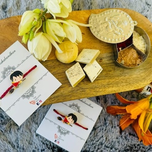 May include: A wooden serving tray holds a Rakshabandhan gift set. The set includes two red and gold bracelets with a cartoon character, sweets, and a decorative container. The cards read "Happy Rakshabandhan". Orange and white flowers are arranged around the tray.