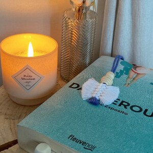 May include: A white candle with a pink label that reads "La Petite Attention" is lit and sitting on a wooden surface. A book with a blue cover and the text "Dangeroux" is on the surface. A white and blue tassel bookmark is on the book.