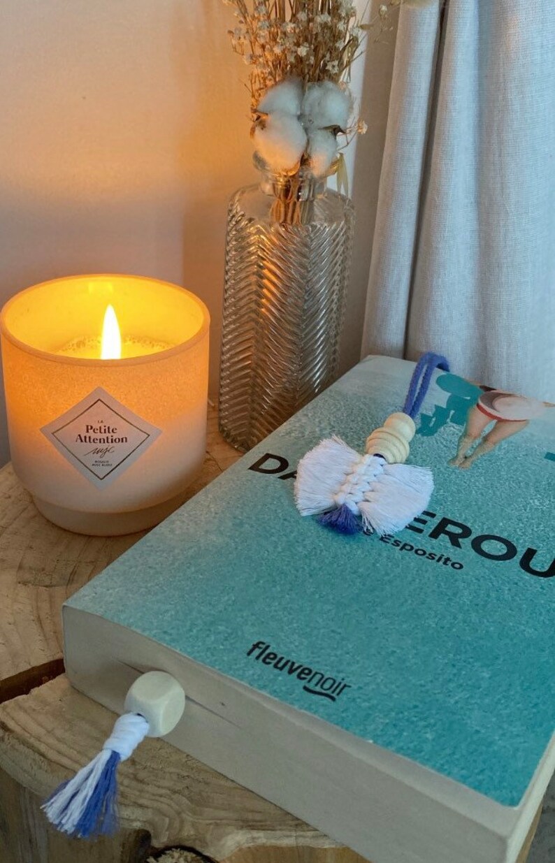 May include: A white candle with a pink label that reads "La Petite Attention" is lit and sitting on a wooden surface. A book with a blue cover and the text "Dangeroux" is on the surface. A white and blue tassel bookmark is on the book.
