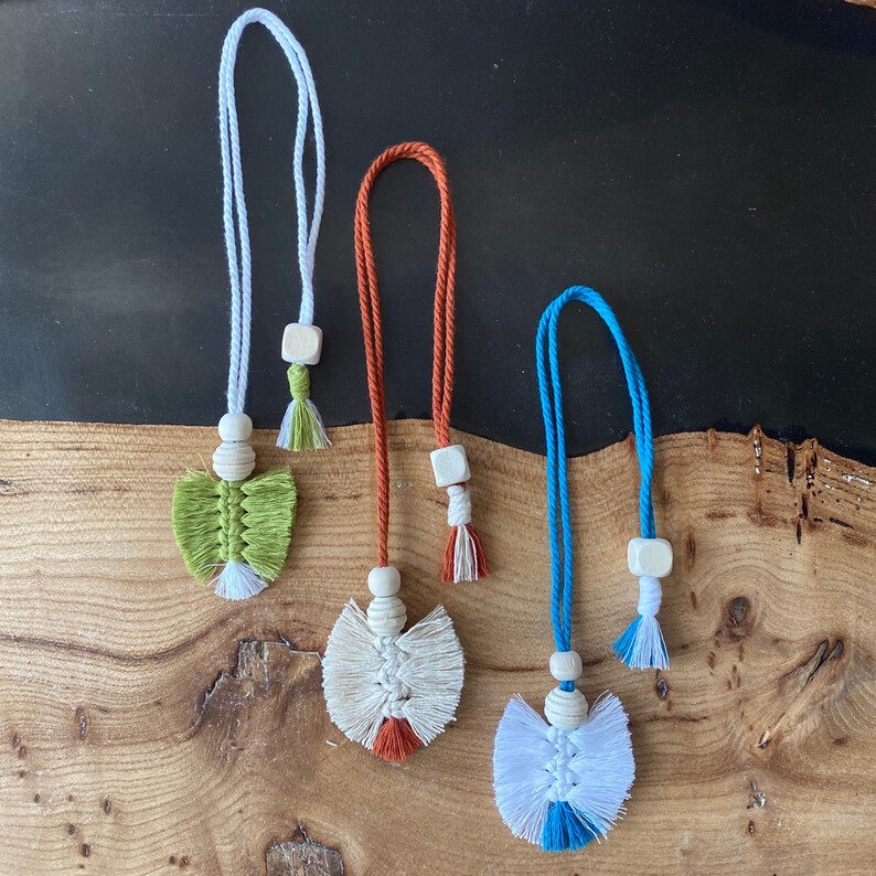 May include: Three handmade macrame tassel keychains with wooden beads and different colored tassels. The tassels are green, white, and blue.
