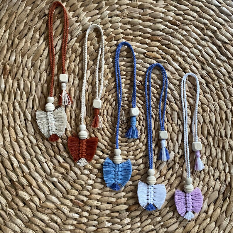 May include: Seven handmade macrame tassel keychains with wooden beads and different colored tassels. The tassels are in shades of white, blue, pink, and brown. The keychains are hanging on a woven straw background.