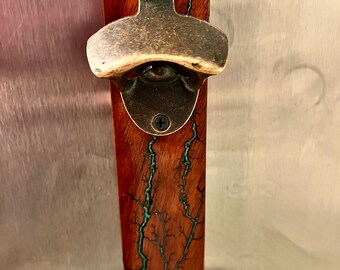 Puede incluir: Un abridor de botellas de madera con un abridor de tapas de metal. La madera tiene una mancha marrón oscura con resina epoxi azul y verde que fluye a través de ella.