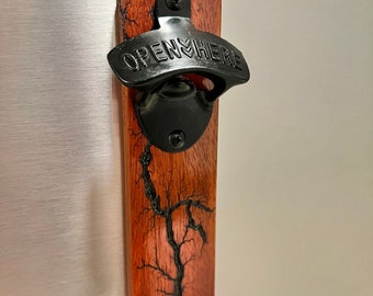 Puede incluir: Un abridor de botellas de madera con un abridor de metal negro y las palabras "OPEN HERE" grabadas en él. La madera tiene un diseño de resina negra.