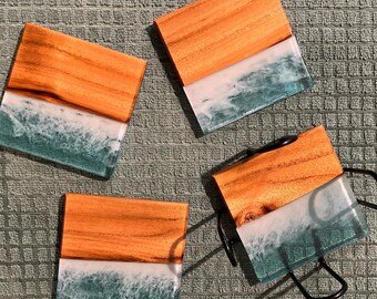 Puede incluir: Cuatro posavasos cuadrados con una parte superior de vetas de madera y una base de resina. La resina es blanca y verde azulado, parecida a las olas del océano. Los posavasos están colocados sobre una superficie verde texturizada.