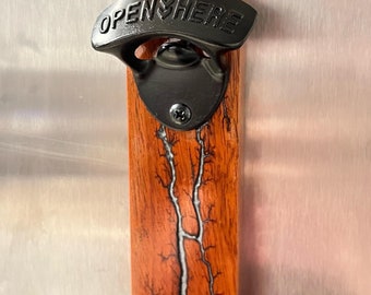 Puede incluir: Un abridor de botellas de metal negro montado en una placa de madera con un diseño de rayo rojo y blanco. El abridor de botellas tiene las palabras "OPEN HERE" en relieve.