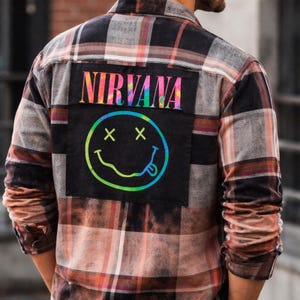 May include: A long-sleeve plaid flannel shirt in shades of brown, black, and white. A large black rectangular patch is sewn on the back, featuring the word NIRVANA in a rainbow gradient font above a rainbow gradient smiley face with X eyes.
