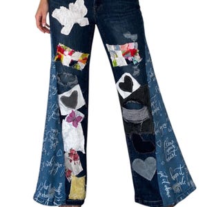 May include: Dark blue denim jeans featuring a wide-leg silhouette, distressed areas, and various patches including white lace, floral fabric, and black hearts. The side panels display white cursive script text, and the pockets have pink stitching.
